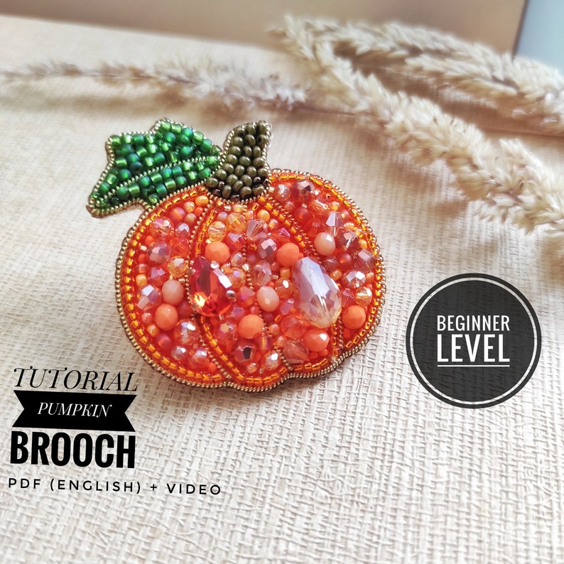 Puede incluir: Una broche de calabaza de cuentas en tonos naranja, verde y dorado. La broche es un proyecto de artesan&iacute;a de nivel principiante. El texto "TUTORIAL PUMPKIN BROOCH PDF (ENGLISH) + VIDEO" est&aacute; debajo de la broche.