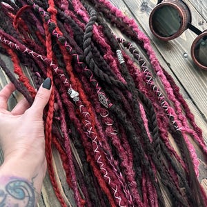 Könnte beinhalten: Handgefertigte Dreadlocks in Rot-, Burgunder- und Schwarztönen. Einige Dreadlocks sind geflochten, andere mit silberfarbenen Charms und weißen Nähten verziert. Eine Steampunk-Brille liegt auf einer Holzoberfläche.