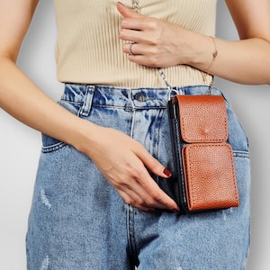 May include: A brown leather crossbody phone bag with a black flap and a silver chain strap. The bag is worn by a person wearing blue jeans.