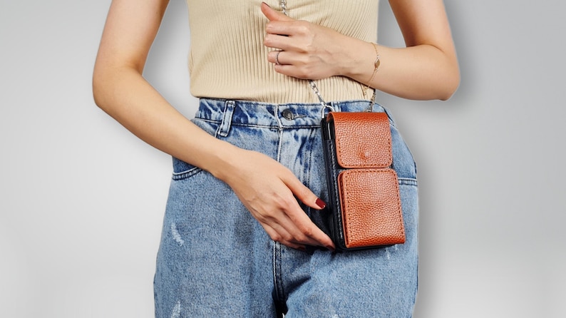 May include: A brown leather crossbody phone bag with a black flap and a silver chain strap. The bag is worn by a person wearing blue jeans.
