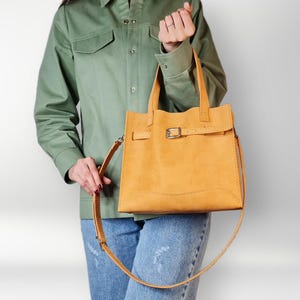 May include: A yellow leather tote bag with a buckle closure and a long strap. The bag is being held by a person wearing a green shirt and blue jeans.