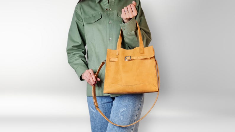 May include: A yellow leather tote bag with a buckle closure and a long strap. The bag is being held by a person wearing a green shirt and blue jeans.