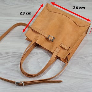 May include: A tan leather tote bag with a buckle closure and a long adjustable strap. The bag measures 26 cm wide, 23 cm tall, and 10 cm deep.