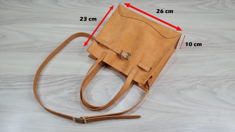 May include: A tan leather tote bag with a buckle closure and a long adjustable strap. The bag measures 26 cm wide, 23 cm tall, and 10 cm deep.