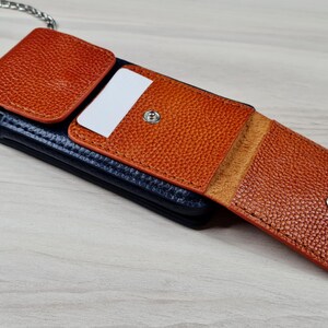 May include: A brown leather phone case with a chain strap. The case has a flap that folds over the phone and snaps closed. The case has a pocket on the front for cards.