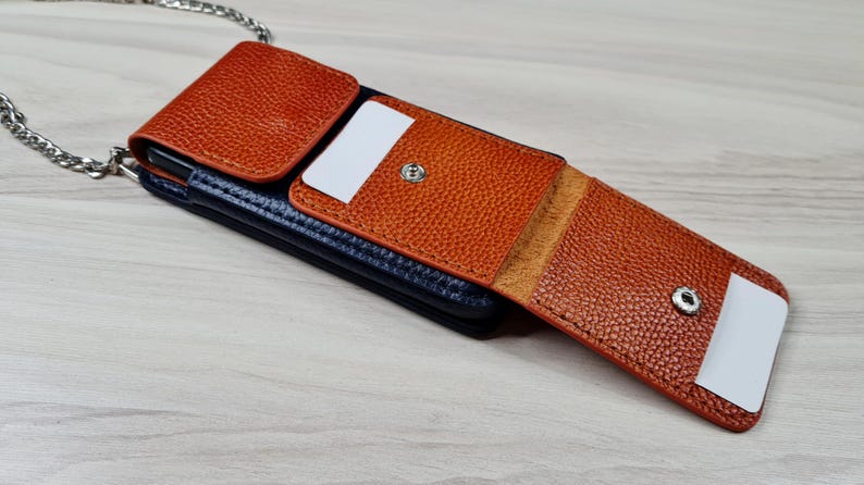May include: A brown leather phone case with a chain strap. The case has a flap that folds over the phone and snaps closed. The case has a pocket on the front for cards.