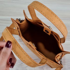 May include: A tan leather handbag with a top handle and a shoulder strap. The bag has a buckle closure and is open to reveal the interior.
