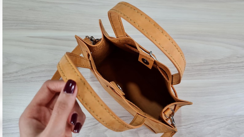 May include: A tan leather handbag with a top handle and a shoulder strap. The bag has a buckle closure and is open to reveal the interior.