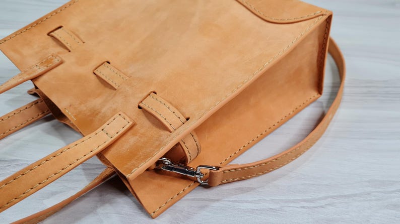 May include: A tan leather tote bag with a top handle and a shoulder strap. The bag has a flap closure and a decorative stitching detail.