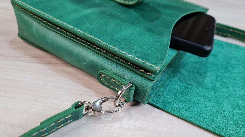 Może przedstawiać: Zielona sk&oacute;rzana torba na telefon na ramię z klapką i srebrnym zapięciem. Torba ma długi pasek, a telefon jest częściowo widoczny w środku. Szwy są białe, a sk&oacute;ra ma teksturowany wygląd.