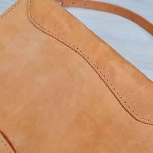 May include: A tan leather crossbody bag with a flap closure and a long strap. The bag has a stitched design and a worn look.