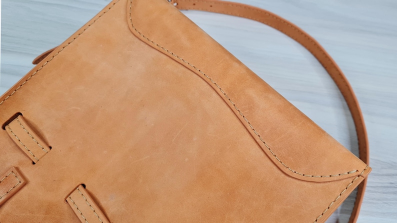 May include: A tan leather crossbody bag with a flap closure and a long strap. The bag has a stitched design and a worn look.