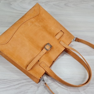 May include: A tan leather tote bag with a buckle closure and a long strap. The bag has a top handle and a shoulder strap.