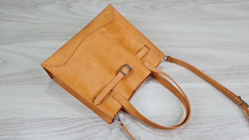 May include: A tan leather tote bag with a buckle closure and a long strap. The bag has a top handle and a shoulder strap.