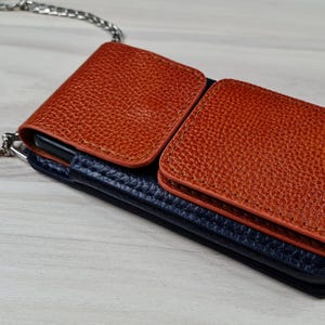 May include: A brown leather phone case with a blue leather flap and a silver chain strap. The case has two pockets on the front.