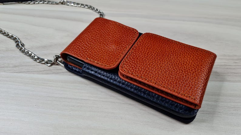 May include: A brown leather phone case with a blue leather flap and a silver chain strap. The case has two pockets on the front.