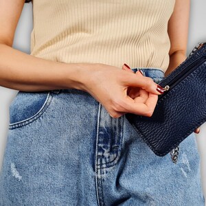 May include: A woman wearing a light beige tank top and blue denim jeans is holding a small black leather crossbody bag with a silver chain strap.