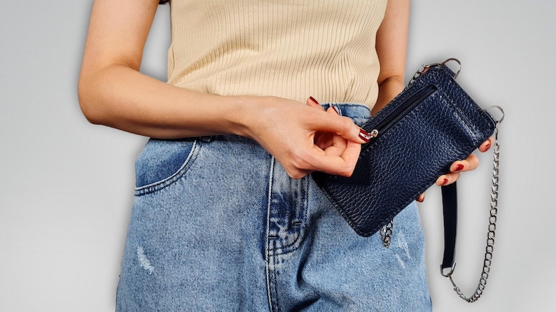 May include: A woman wearing a light beige tank top and blue denim jeans is holding a small black leather crossbody bag with a silver chain strap.