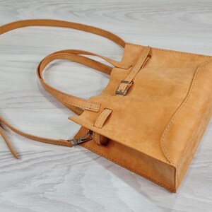 May include: A light brown leather tote bag with a buckle closure and a long strap. The bag has a top handle and a shoulder strap.