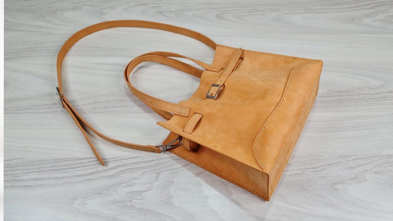 May include: A light brown leather tote bag with a buckle closure and a long strap. The bag has a top handle and a shoulder strap.
