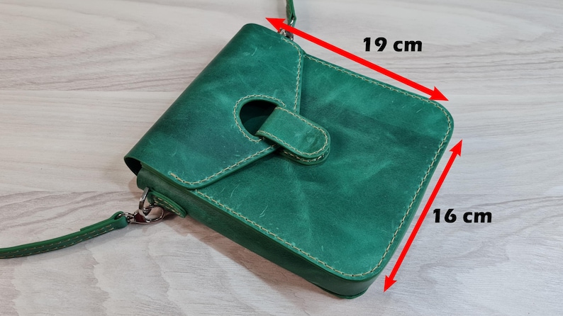 Może przedstawiać: Zielona sk&oacute;rzana torba na ramię z klapką i paskiem. Torba ma około 19 cm szerokości i 16 cm wysokości. Posiada okrągły otw&oacute;r na klapie i białe przeszycia. Torba wykonana jest z teksturowanej sk&oacute;ry.