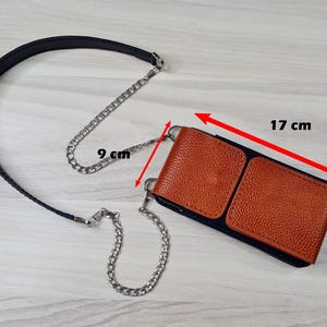 May include: A brown leather phone case with a black strap and a silver chain. The case measures 17 cm by 9 cm.