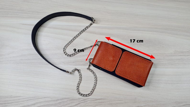 May include: A brown leather phone case with a black strap and a silver chain. The case measures 17 cm by 9 cm.