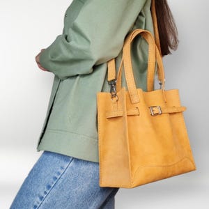 May include: A yellow leather tote bag with a buckle closure and a long strap. The bag is being carried by a person wearing a green jacket and blue jeans.