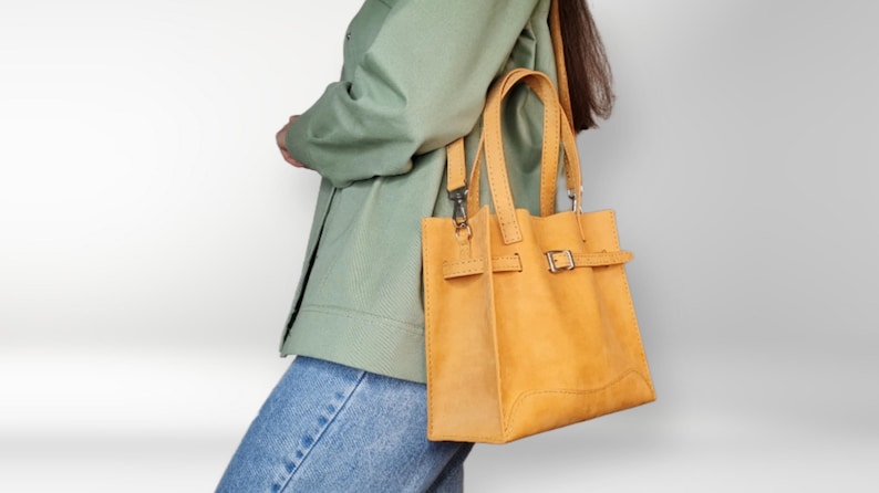 May include: A yellow leather tote bag with a buckle closure and a long strap. The bag is being carried by a person wearing a green jacket and blue jeans.