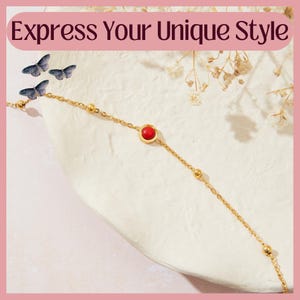 Pode incluir: Colar dourado com uma pedra vermelha central. A corrente é delicada, com pequenas contas douradas. A imagem inclui o texto "Express Your Unique Style" em uma faixa rosa, juntamente com borboletas e detalhes florais.