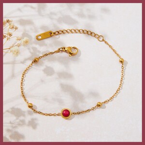 May include: Gold-toned bracelet featuring a small, round, red gemstone at the center. The bracelet has a delicate chain design with small, evenly spaced gold beads. The clasp and connecting elements are also gold. The background is a soft, neutral color.