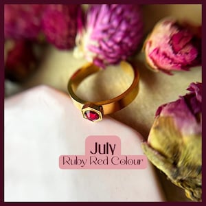 May include: A gold ring with a ruby red gemstone set in a hexagonal shape. The ring is on a white surface with dried pink flowers in the background. The text "July Ruby Red Colour" is below the ring.