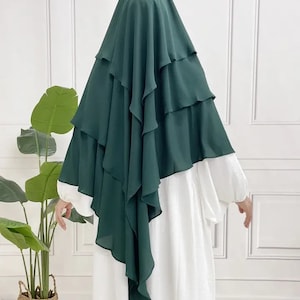 May include: A dark green layered chiffon hijab with a ruffled edge. The hijab is draped over the shoulders and head, with the ends flowing down the back.