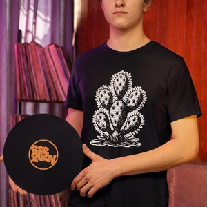 Może przedstawiać: Czarny t-shirt z białym wzorem kaktusa. Czarna płyta winylowa z pomarańczowym tekstem jest trzymana przed koszulką. Płyta ma tekst "esc Quely" w okrągłym wzorze. Koszulka jest z krótkim rękawem.