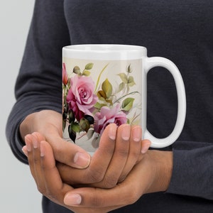 May include: White ceramic mug with a floral design featuring pink roses and green leaves. The mug has a white handle.