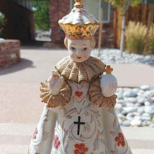 May include: A porcelain figurine of a young child wearing a gold crown and a white robe with floral designs. The child is holding a heart and a cross.