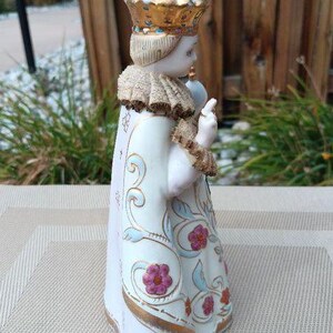 May include: A porcelain figurine of a child wearing a gold and white crown and a white robe with gold and floral designs. The figurine is holding a small object in its right hand.