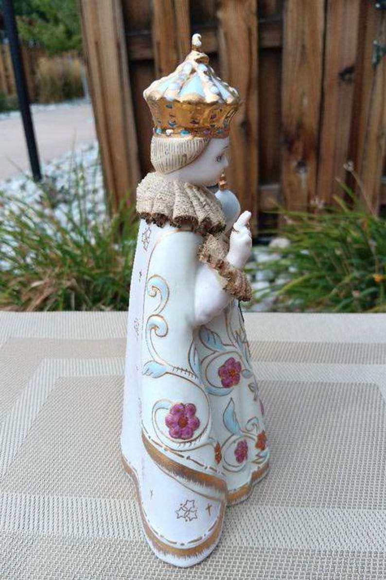 May include: A porcelain figurine of a child wearing a gold and white crown and a white robe with gold and floral designs. The figurine is holding a small object in its right hand.