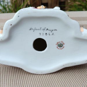 May include: The underside of a white porcelain figurine with the words "Royal Art of Prague" and "7101A" written in black ink. The figurine also has a small red and gold sticker with the words "Saxon China Hand Painted" on it.