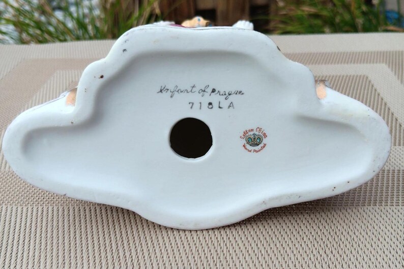 May include: The underside of a white porcelain figurine with the words "Royal Art of Prague" and "7101A" written in black ink. The figurine also has a small red and gold sticker with the words "Saxon China Hand Painted" on it.