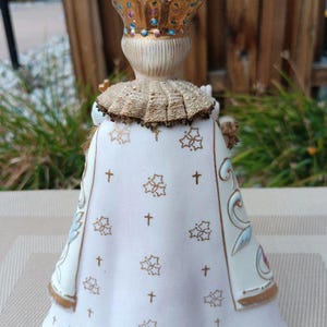 May include: A white porcelain figurine of a child wearing a gold crown and a white robe with gold trim and a pattern of gold crosses and flowers. The figurine is standing on a beige surface.