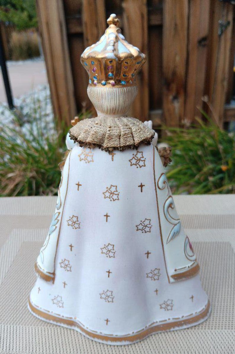 May include: A white porcelain figurine of a child wearing a gold crown and a white robe with gold trim and a pattern of gold crosses and flowers. The figurine is standing on a beige surface.