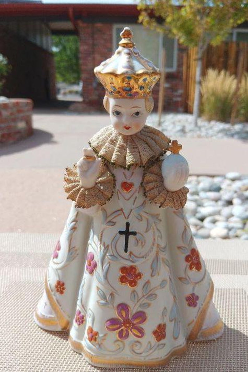 May include: A porcelain figurine of a young child wearing a gold crown and a white robe with floral designs. The child is holding a heart and a cross.