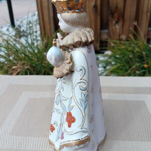 May include: A porcelain figurine of a young person wearing a white and gold robe with floral designs. The figurine is wearing a gold crown with blue and white accents. The figurine is holding a small white object in their right hand.
