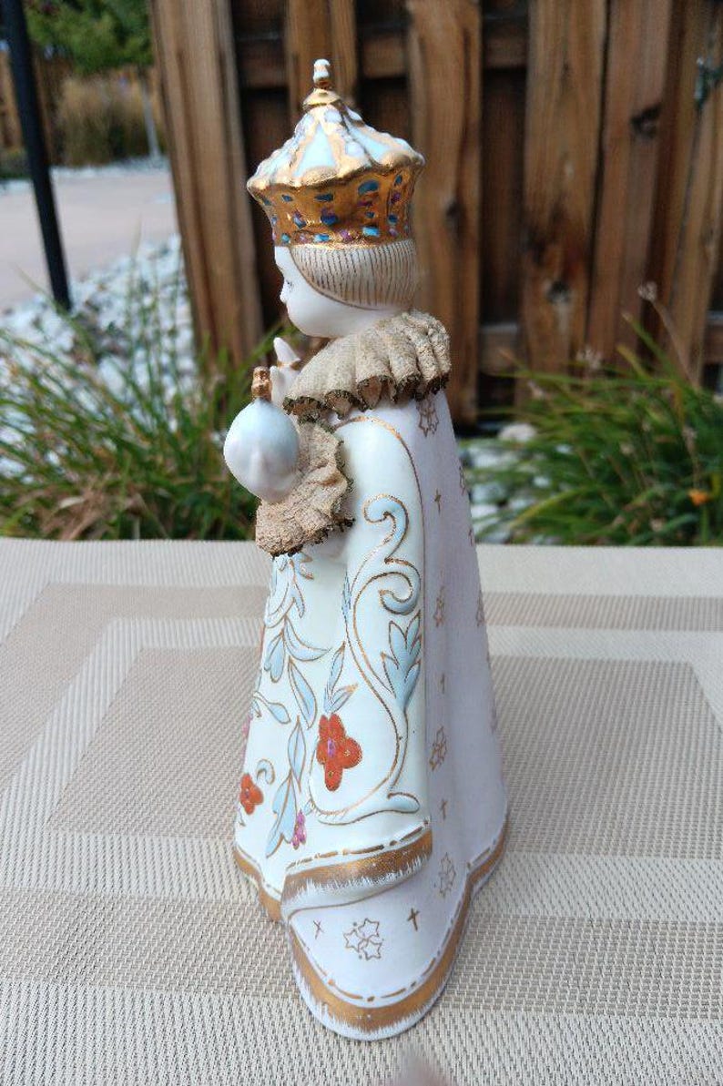 May include: A porcelain figurine of a young person wearing a white and gold robe with floral designs. The figurine is wearing a gold crown with blue and white accents. The figurine is holding a small white object in their right hand.