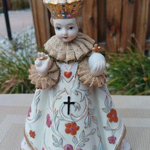 May include: A porcelain figurine of a child wearing a white dress with gold trim and floral designs. The child is wearing a gold crown and holding a small white orb in their right hand. The figurine is standing on a white surface.