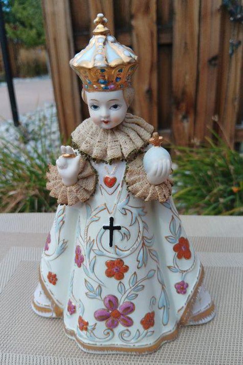 May include: A porcelain figurine of a child wearing a white dress with gold trim and floral designs. The child is wearing a gold crown and holding a small white orb in their right hand. The figurine is standing on a white surface.