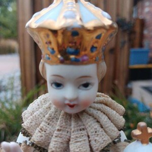 May include: A porcelain figurine of a young child wearing a gold and white crown and a cream-coloured lace collar. The figurine is holding a small golden object in its right hand.
