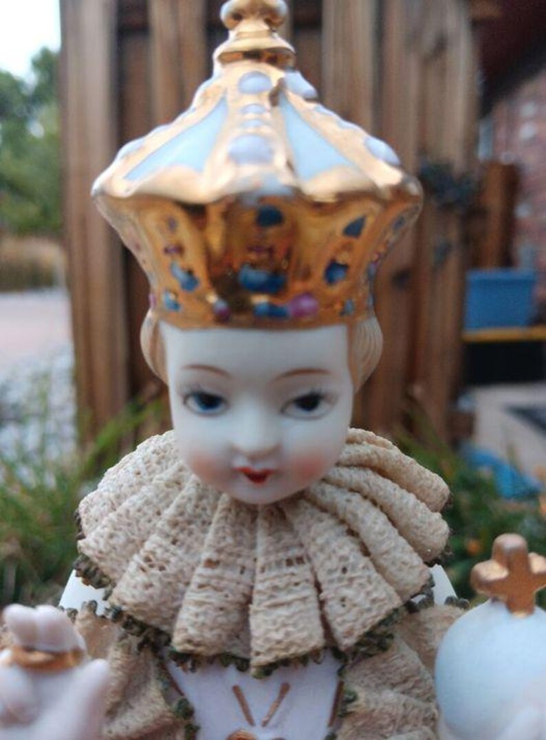 May include: A porcelain figurine of a young child wearing a gold and white crown and a cream-coloured lace collar. The figurine is holding a small golden object in its right hand.