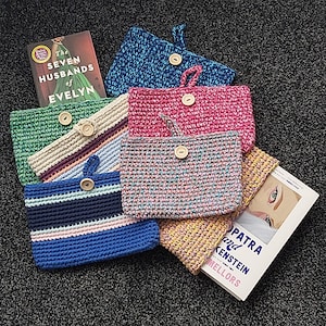 May include: A stack of seven crocheted book covers in various colours, including green, blue, pink, and beige. The covers are decorated with buttons and have a textured, woven appearance. The book covers are stacked on top of a book titled "The Seven Husbands of Evelyn".